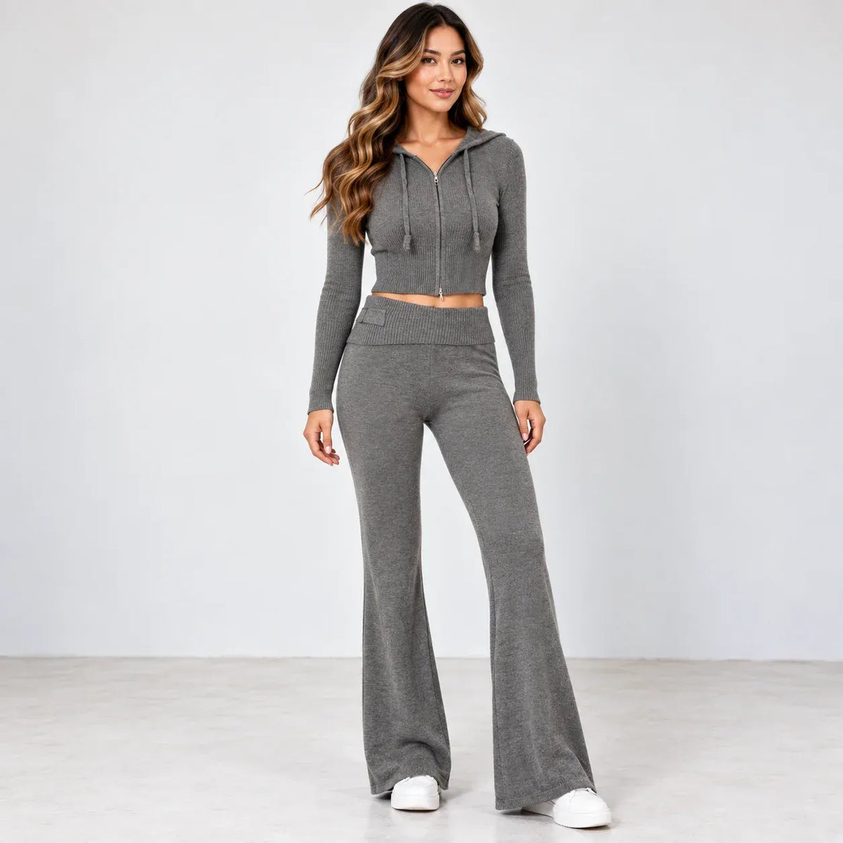 A woman modeling a matching gray two-piece knit loungewear set, featuring a cropped zip-up hoodie with drawstrings and high-waisted, wide-leg flare pants, paired with white sneakers against a light gray studio background.
