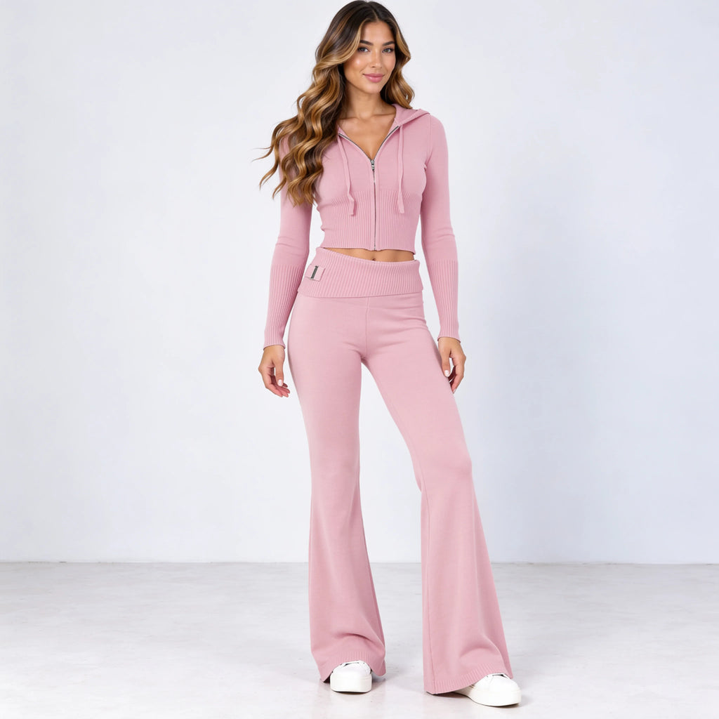 A woman modeling a matching dusty pink two-piece knit set consisting of a cropped zip-up hoodie and high-waisted flared pants, paired with white sneakers against a light gray studio background.