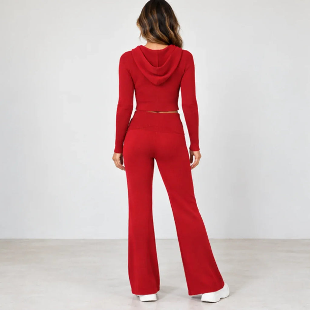A woman viewed from behind wearing a bright red two-piece outfit consisting of a long-sleeve cropped hoodie with a small waist cutout and matching high-waisted flared pants, against a plain light gray studio background.