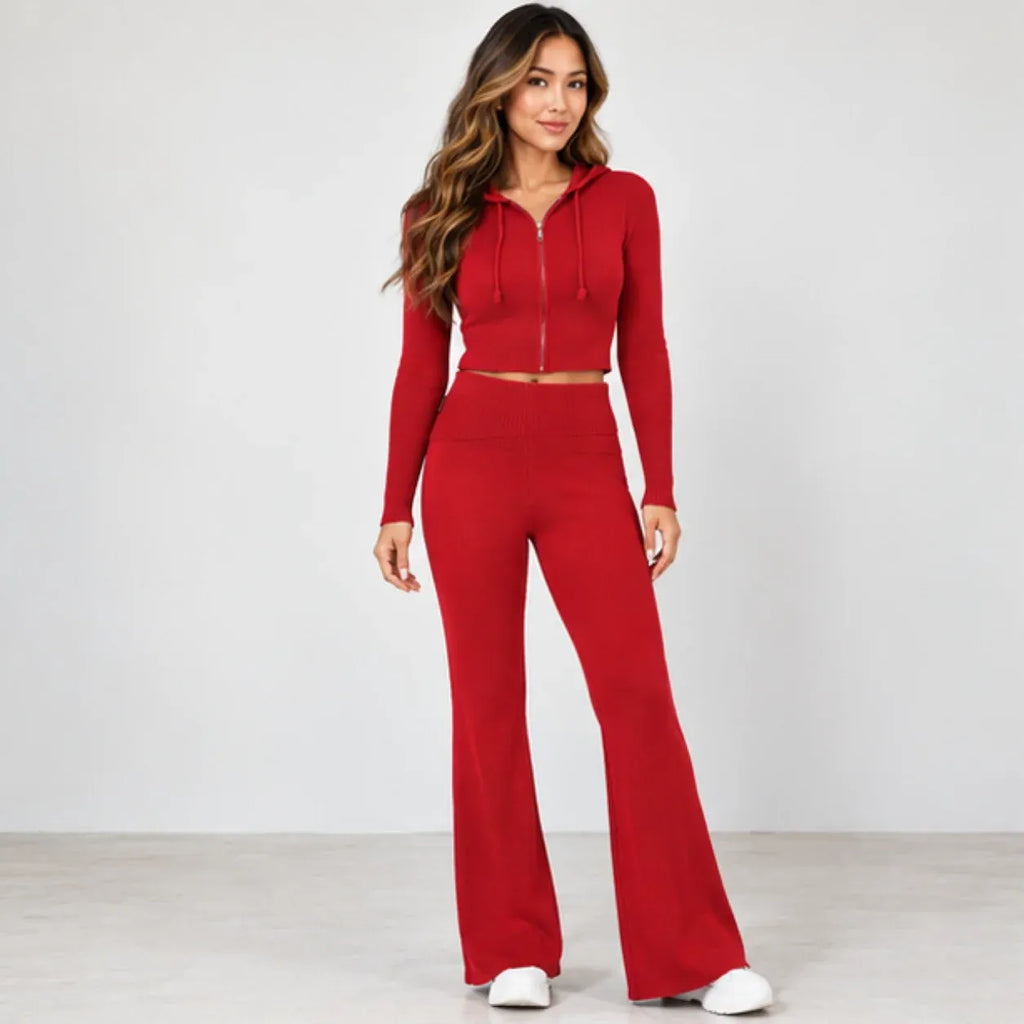 A woman modeling a matching red two-piece set consisting of a cropped zip-up hoodie with drawstrings and high-waisted flared pants, photographed against a plain light gray studio background.
