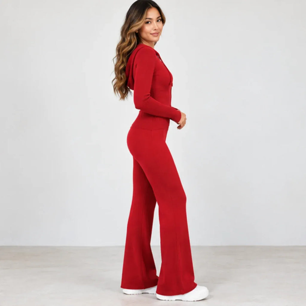 A woman modeling a bright red long-sleeve jumpsuit with a high neckline and wide-leg flared pants, paired with white sneakers, against a plain light gray studio background.