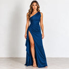 A woman modeling a floor-length one-shoulder gown in deep blue satin, featuring a high thigh slit and draped skirt, set against a plain white studio background.