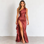 A woman models a floor-length one-shoulder satin gown in a dusty rose or mauve shade, featuring a high thigh slit and draped skirt, set against a plain light gray studio background.