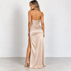 A woman modeling a floor-length satin gown from the back, featuring thin spaghetti straps, a low open back, and a high thigh slit. The dress is a champagne beige color with a satin sheen, paired with matching beige high-heeled sandals, set against a plain light gray studio background.