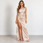 A woman modeling a floor-length, one-shoulder satin gown in a champagne beige color, standing against a neutral light gray studio background.