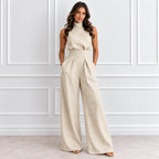 A woman modeling a sleeveless high-neck beige jumpsuit with wide legs and a matching fabric belt, standing against a white paneled wall.