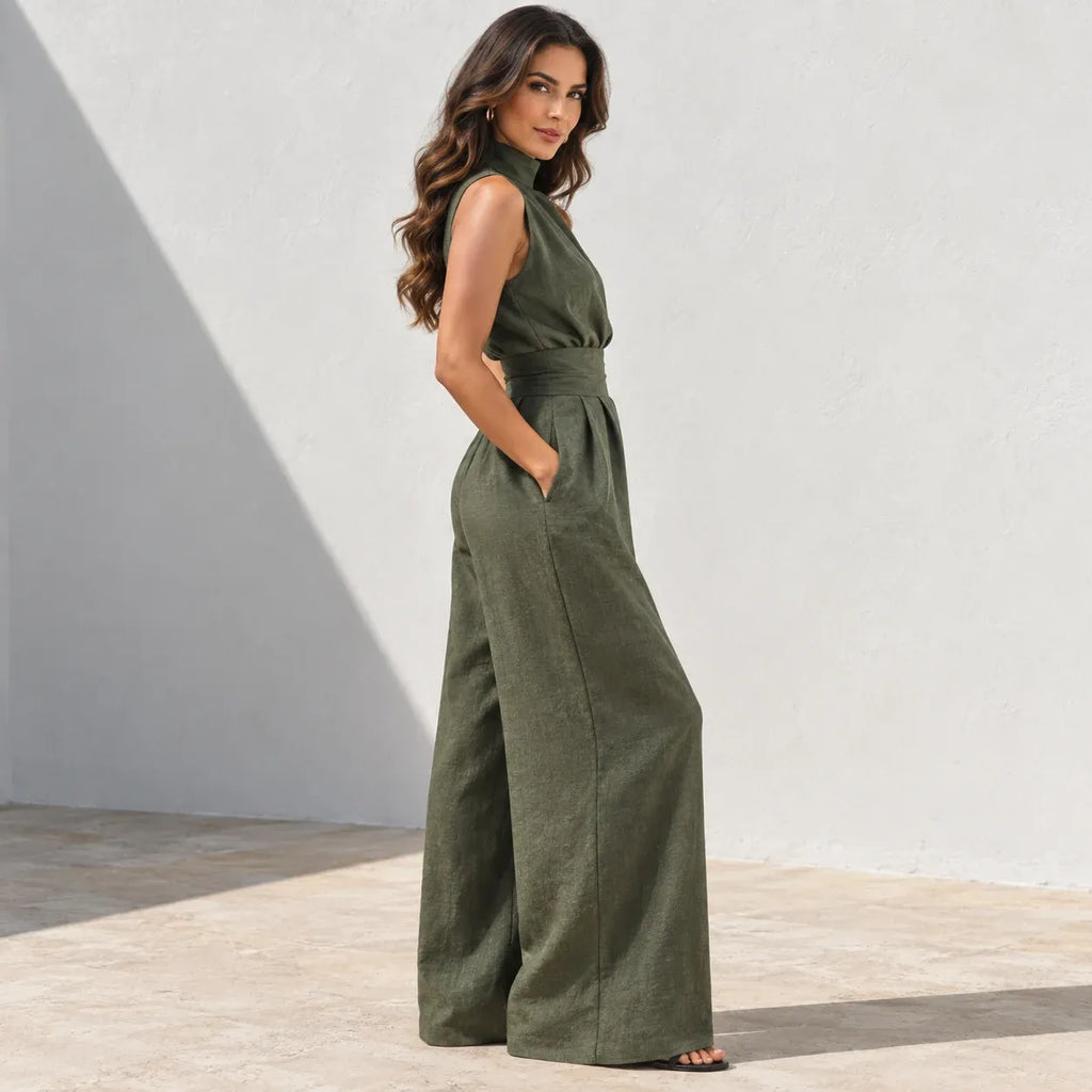 A woman modeling an olive green sleeveless wide-leg jumpsuit in a minimalist outdoor setting with a textured white wall and concrete floor.