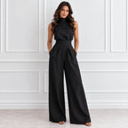 A woman modeling a black sleeveless high-neck wide-leg jumpsuit with a matching waistband, standing against a white paneled wall.