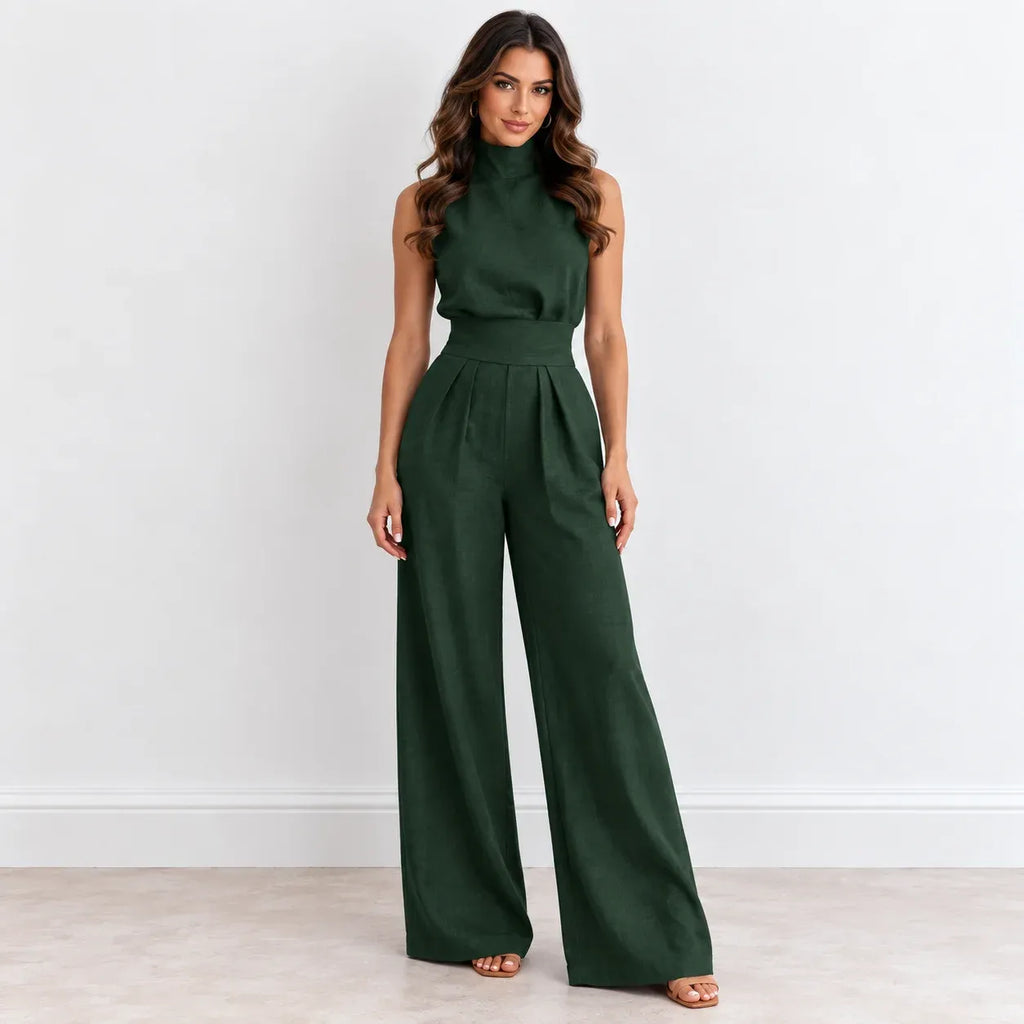 A woman modeling a dark forest green sleeveless jumpsuit with a mock neck, defined waist, and wide-leg pants, standing against a plain white wall.