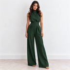 A woman modeling a dark forest green sleeveless jumpsuit with a mock neck, defined waist, and wide-leg pants, standing against a plain white wall.