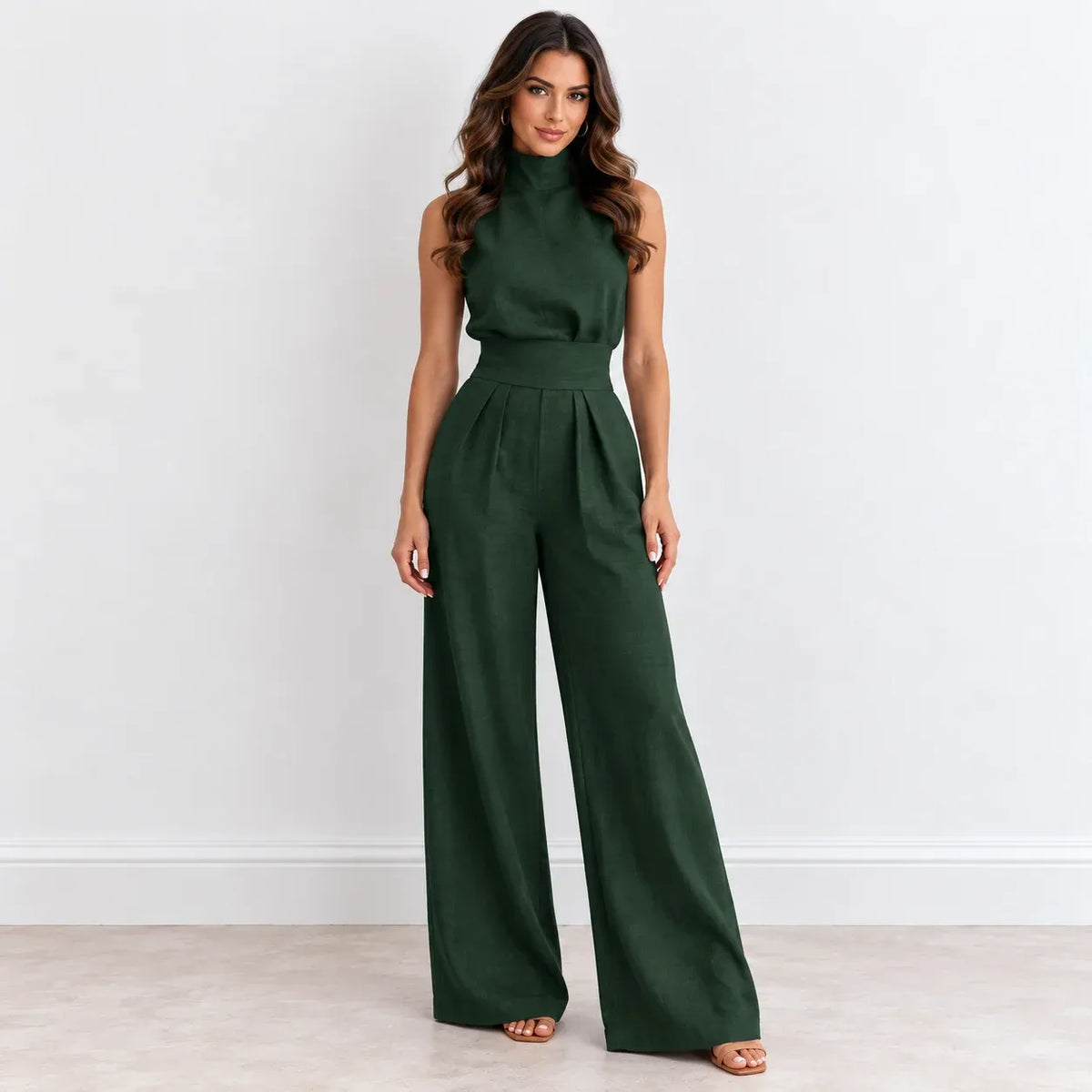 A woman modeling a dark forest green sleeveless jumpsuit with a mock neck, defined waist, and wide-leg pants, standing against a plain white wall.