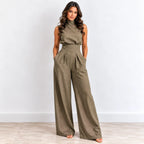 A woman modeling a sleeveless high-waisted olive green jumpsuit with wide legs, standing against a light gray wall and light wood floor.
