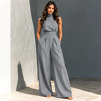 A woman modeling a gray sleeveless high-neck jumpsuit with wide-leg pleated pants, standing in a sunlit modern concrete setting.