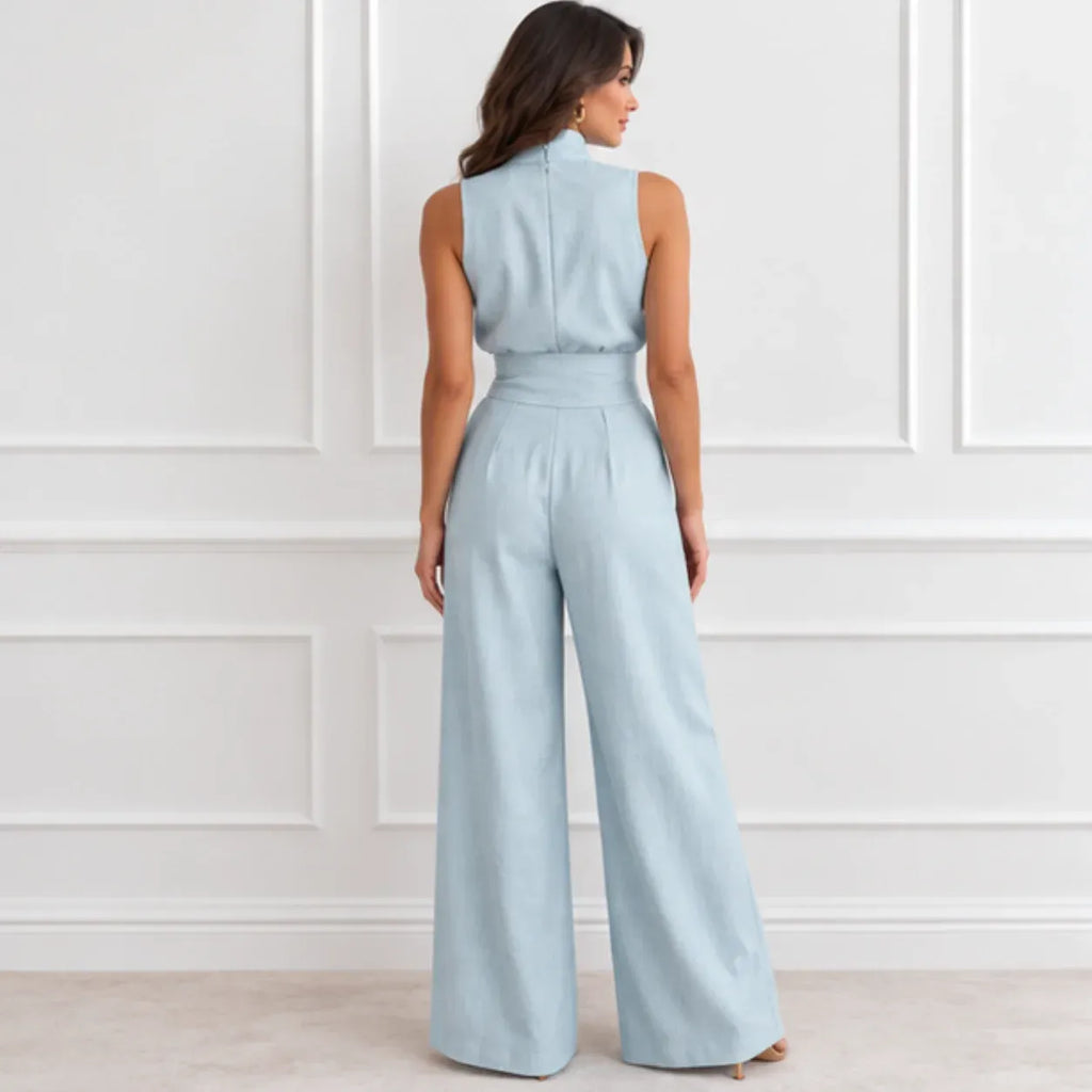 A woman viewed from the back wearing a light blue sleeveless jumpsuit with wide legs and a defined waist, standing against a white paneled wall.