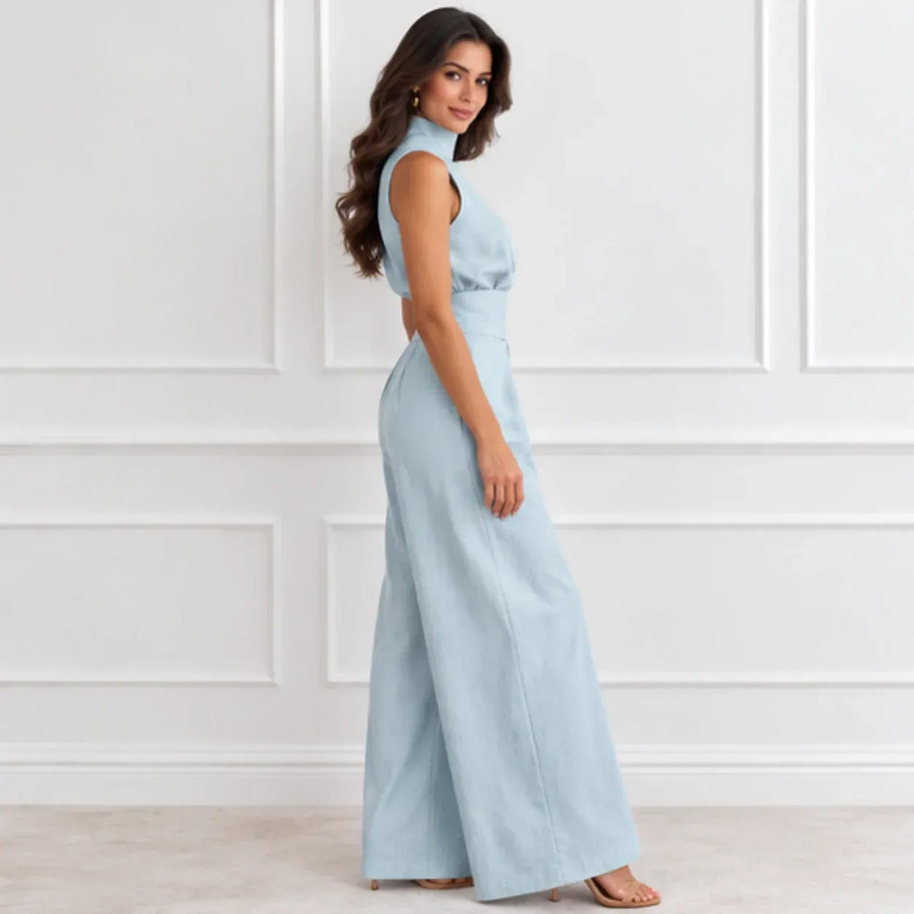 A woman modeling a light blue sleeveless maxi dress with a high mock neck, side cutout, and wide-leg pants, standing against a white paneled wall.