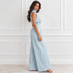 A woman modeling a light blue sleeveless maxi dress with a high mock neck, side cutout, and wide-leg pants, standing against a white paneled wall.