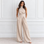 A woman modeling a sleeveless high-neck beige jumpsuit with wide legs, standing against a white paneled wall.