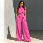 A woman models a bright pink sleeveless high-neck jumpsuit with wide-leg trousers, standing in a minimalist studio setting with a textured gray wall and tiled floor.