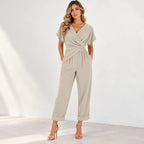 A woman modeling a light beige short-sleeve wrap-style jumpsuit with a twisted waist detail and cuffed hems, paired with nude high-heeled sandals against a bright white studio background.