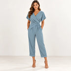 A woman modeling a light blue wrap-style jumpsuit with short sleeves, a V-neckline, side pockets, and cropped cuffed legs, paired with tan heeled sandals against a plain white studio background.