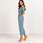 A woman modeling a dusty blue short-sleeve jumpsuit with a cuffed ankle and side pockets, paired with nude high-heeled sandals, against a clean white studio background.