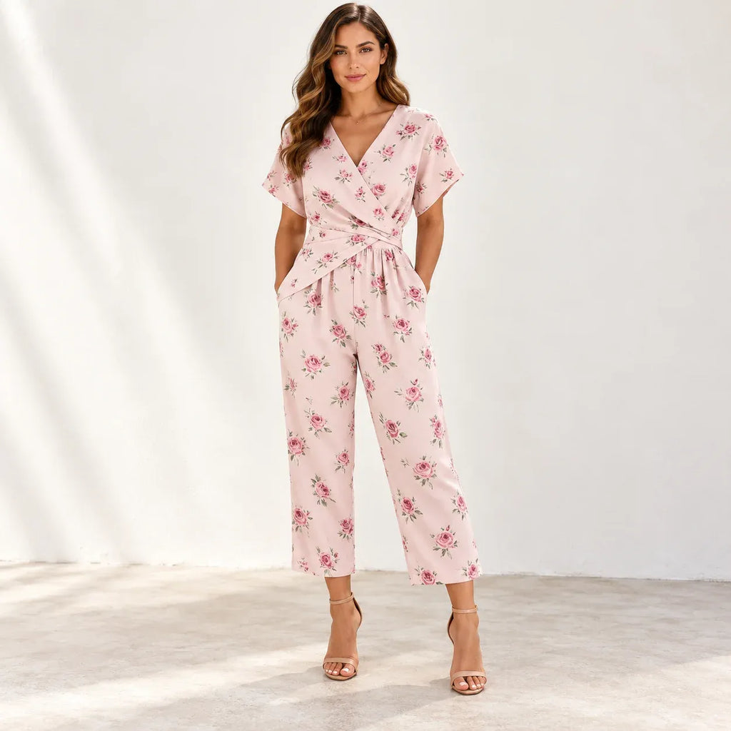 A woman models a light pink wrap-style jumpsuit with a delicate pink and green floral print, featuring short flutter sleeves, a V-neckline, side pockets, and a self-tie waist, paired with tan high-heeled sandals against a bright, sunlit studio backdrop.