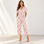A woman models a light pink wrap-style jumpsuit with a delicate pink and green floral print, featuring short flutter sleeves, a V-neckline, side pockets, and a self-tie waist, paired with tan high-heeled sandals against a bright, sunlit studio backdrop.