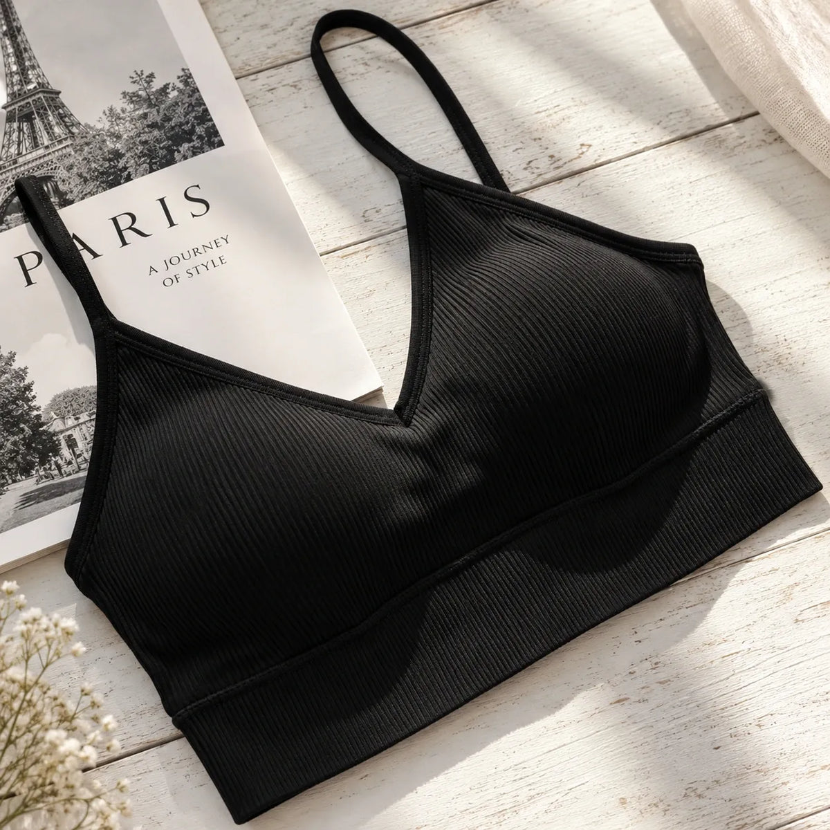 A black ribbed wireless bra with a V-neckline and thin adjustable straps, styled on a white wooden surface next to a Paris magazine and dried flowers.