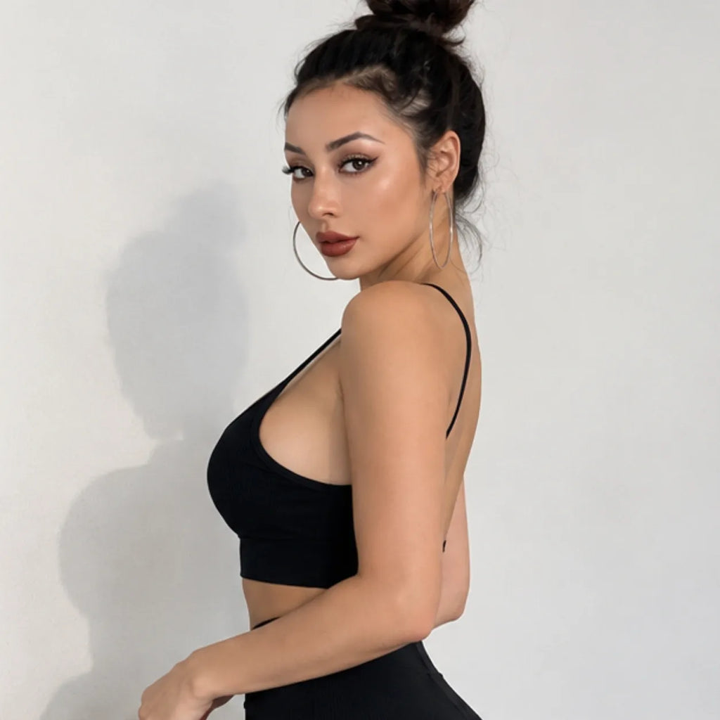 A woman with her hair in a high bun models a black strappy crop top and matching black bottoms against a plain light gray wall.