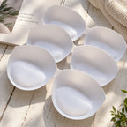 Six white silicone breast pads arranged in a circular pattern on a light wooden surface, with a book, towel, and small plant leaves in the background.