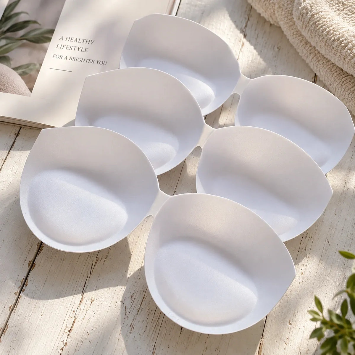 Six white silicone breast pads arranged in a circular pattern on a light wooden surface, with a book, towel, and small plant leaves in the background.
