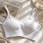 A white ribbed wireless bralette laid flat on a light wood surface with styled props including a magazine, flowers, and a perfume bottle.