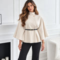 A woman modeling a high-neck, cape-style beige coat with wide sleeves, a single decorative button, and a black belt with gold hardware at the waist, paired with black pants in a bright white interior setting.