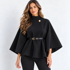 A woman modeling a black high-neck cape-style top with a single gold button and a matching black belt with a gold buckle, paired with black pants against a light gray studio background.