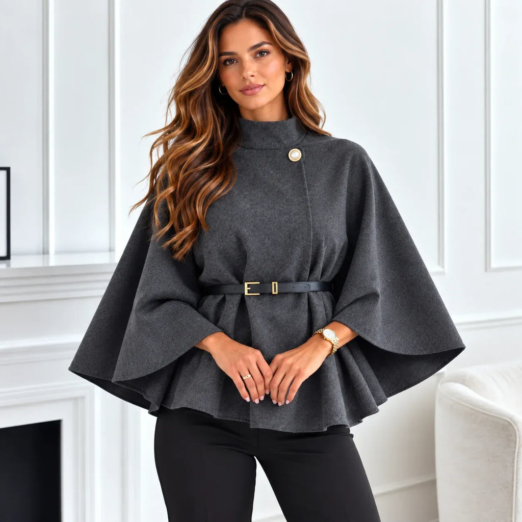 A woman wearing a dark gray cape-style top with a single gold button and a matching dark gray belt with a gold buckle, paired with black pants, standing in a bright white room.