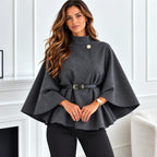 A woman wearing a dark gray cape-style top with a single gold button and a matching dark gray belt with a gold buckle, paired with black pants, standing in a bright white room.