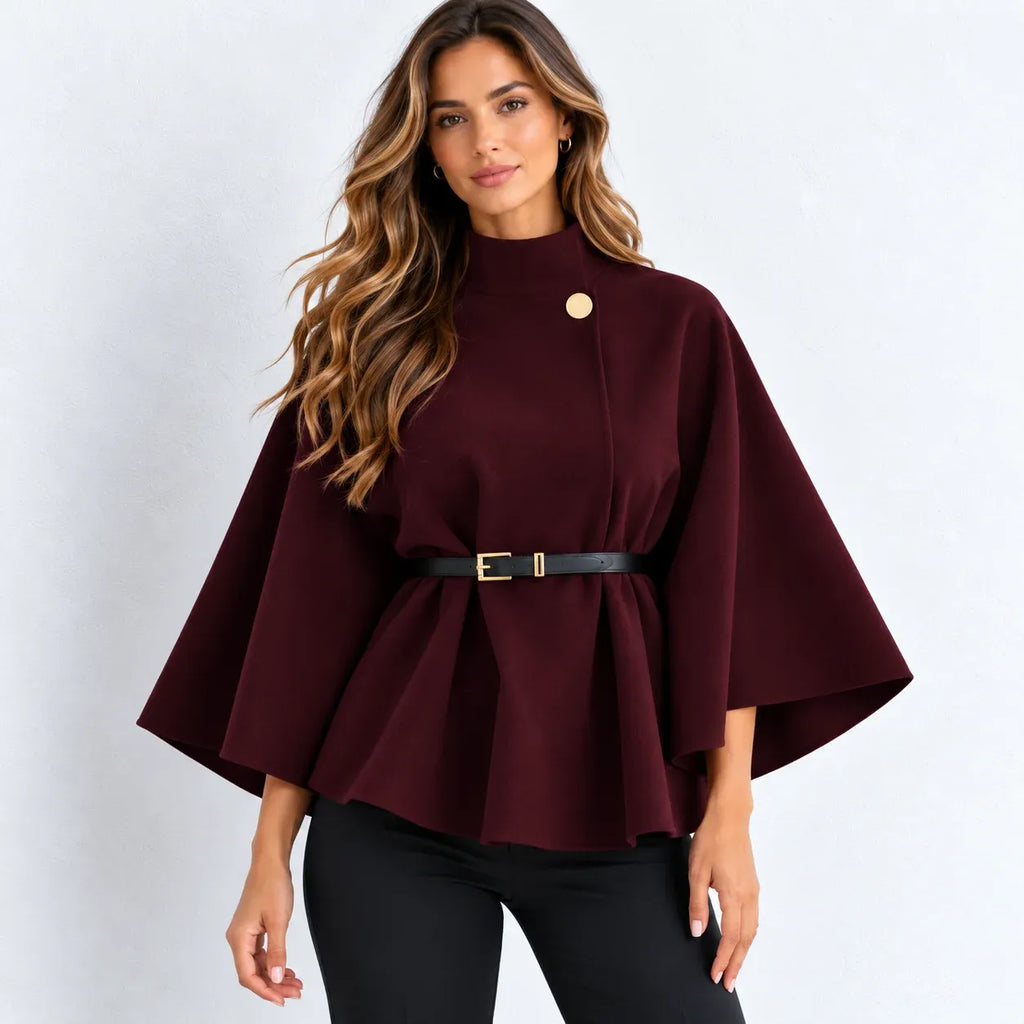 A woman modeling a burgundy cape-style top with a high mock neck, wide sleeves, a peplum hem, a black belt with a gold buckle, and a gold button detail on the chest, paired with black pants against a light gray background.