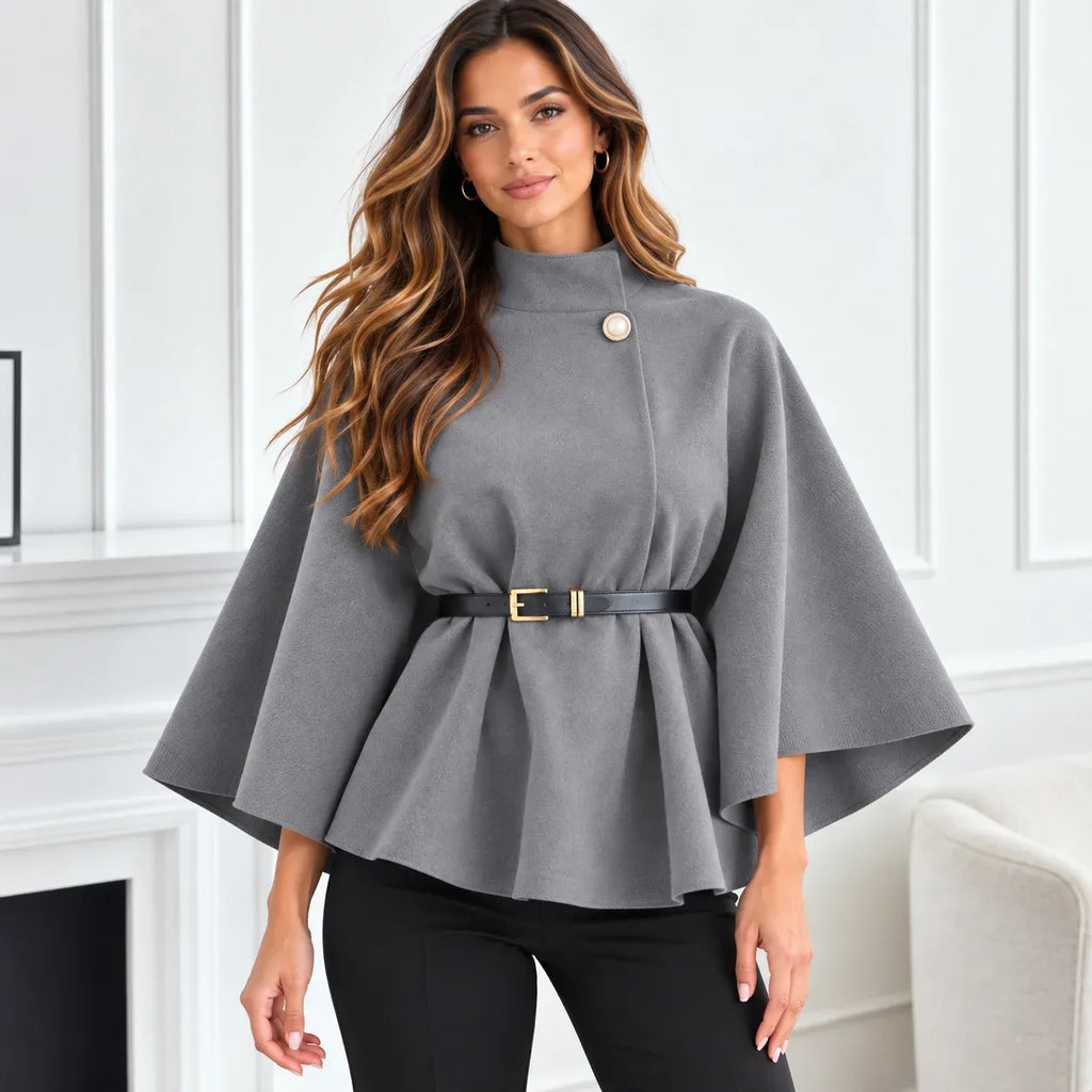 A woman modeling a gray high-neck cape-style top with a single decorative button and a black belt with gold hardware, paired with black pants in a bright indoor setting.