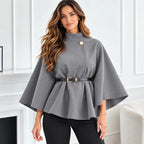 A woman modeling a gray high-neck cape-style top with a single decorative button and a black belt with gold hardware, paired with black pants in a bright indoor setting.