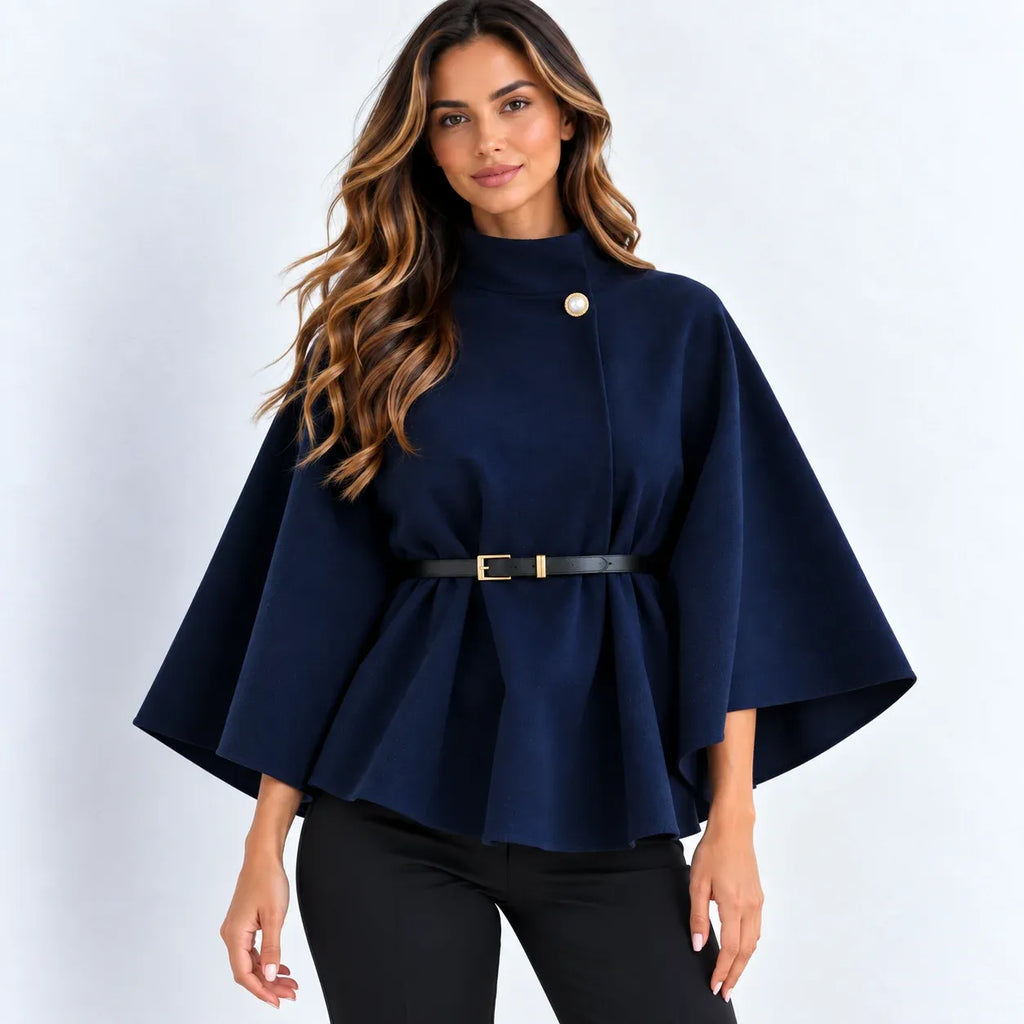A woman modeling a navy blue cape-style top with a single gold-tone button at the collar, a black belt with a gold buckle at the waist, and black pants, set against a plain light gray studio background.