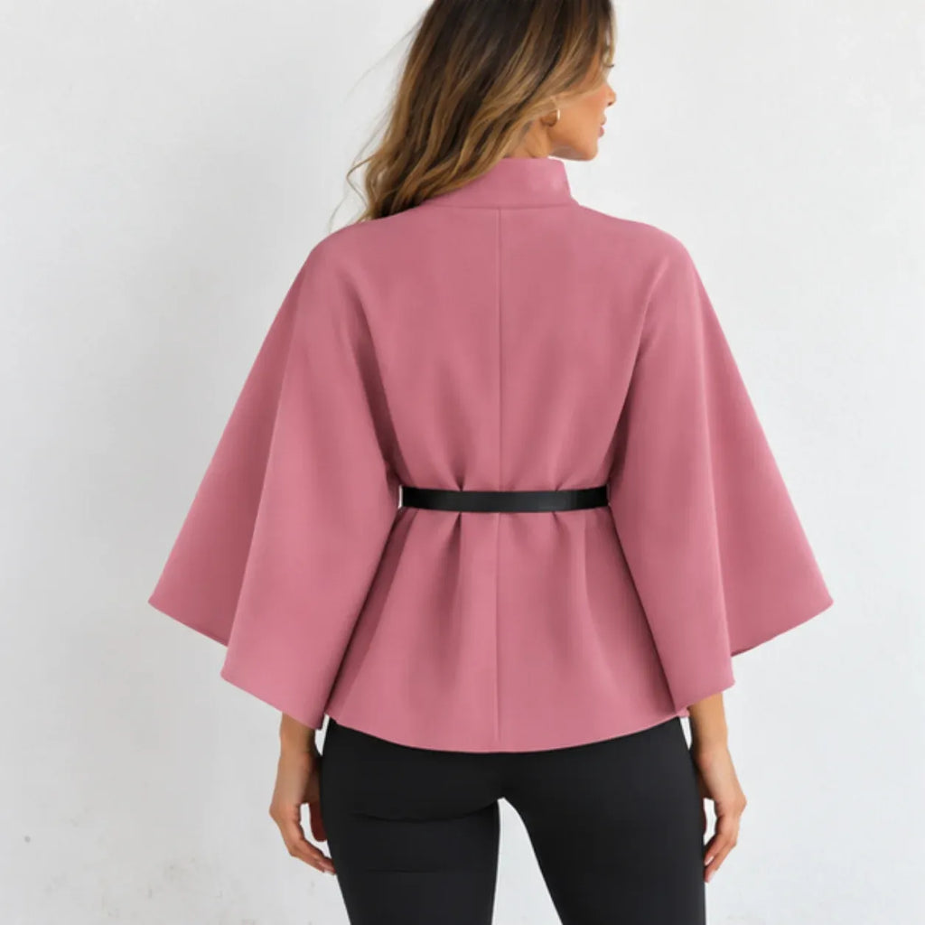 A woman shown from the back wearing a dusty pink cape-style jacket with wide sleeves and a thin black belt at the waist, paired with black pants against a plain white background.