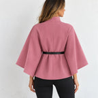 A woman shown from the back wearing a dusty pink cape-style jacket with wide sleeves and a thin black belt at the waist, paired with black pants against a plain white background.