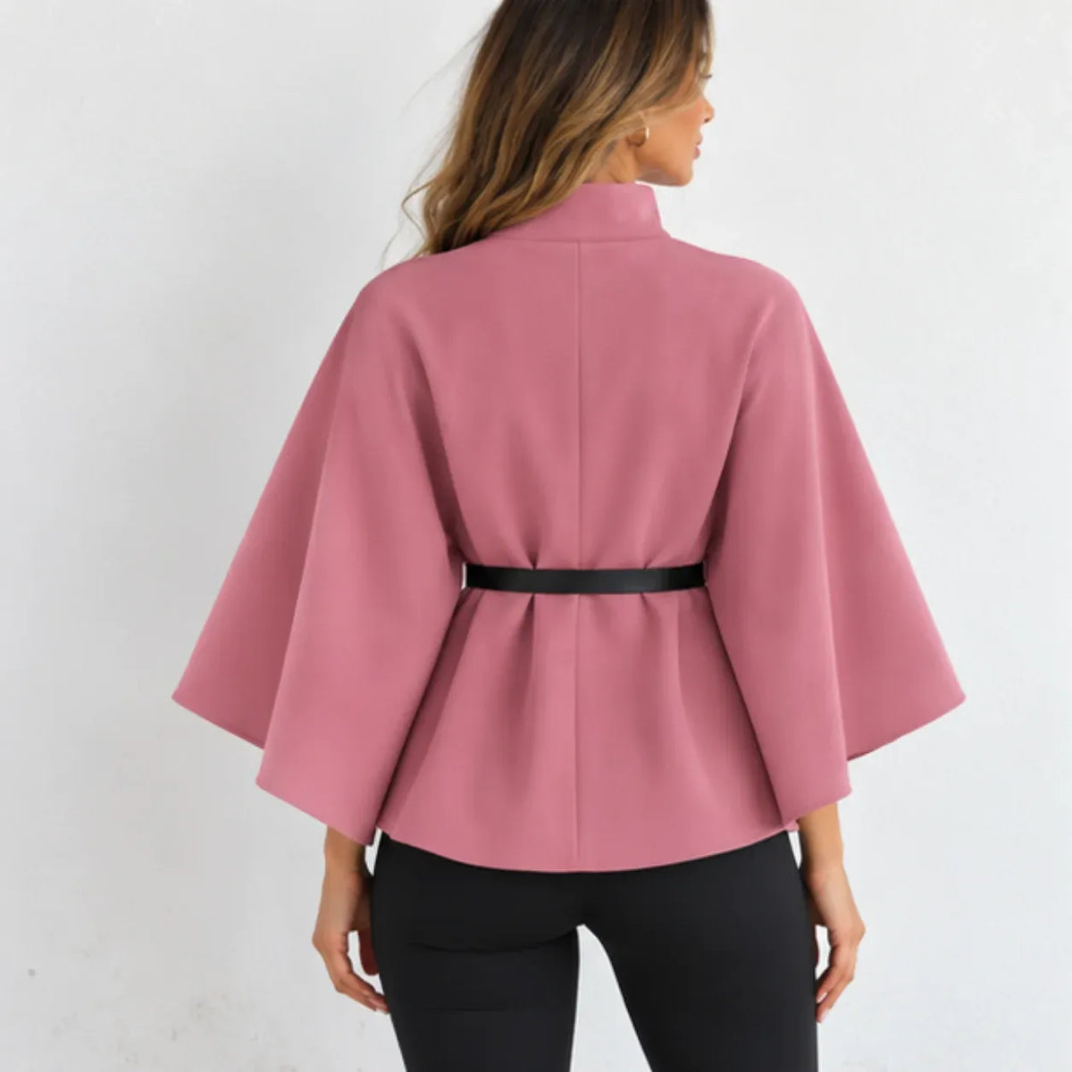 A woman shown from the back wearing a dusty pink cape-style jacket with wide sleeves and a thin black belt at the waist, paired with black pants against a plain white background.