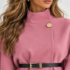 A close-up of a woman wearing a dusty pink high-neck coat or dress with a black belt and gold-tone buckle, featuring a gold button detail on the chest.