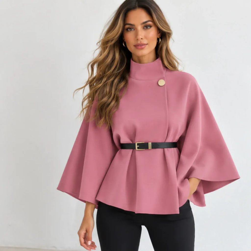 A woman modeling a dusty pink cape-style peplum top with a high mock neck, wide flared sleeves, a black belt, and a gold button accent, paired with black pants against a plain white background.