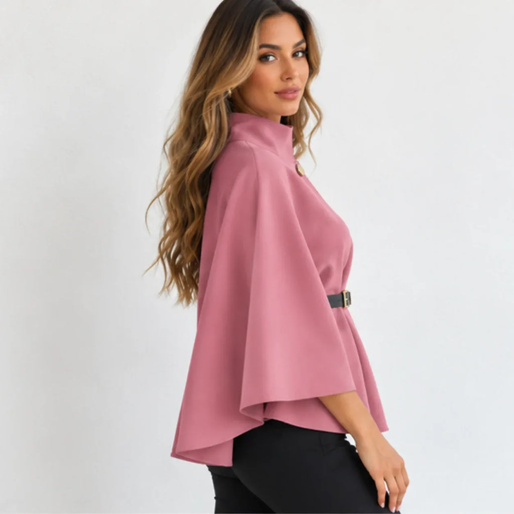 A woman wearing a dusty pink cape-sleeve blouse with a high mock neck and a thin black belt at the waist, paired with black pants, photographed against a light gray studio background.