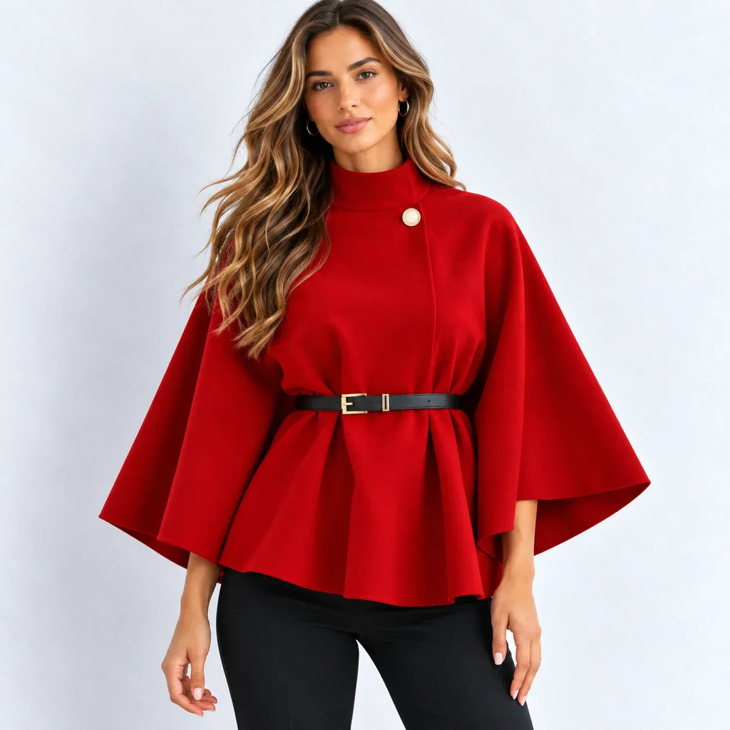 A woman modeling a bright red cape-style peplum top with wide sleeves, a high mock neck, a single decorative button, a black belt with a gold buckle at the waist, and black pants, set against a plain light gray studio background.