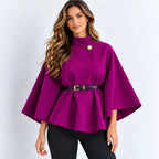 A woman modeling a bright magenta-purple cape-style top with a high mock neck, wide three-quarter sleeves, a peplum hem, a single decorative gold button, and a black belt with a gold buckle at the waist, paired with black pants against a light gray studio background.