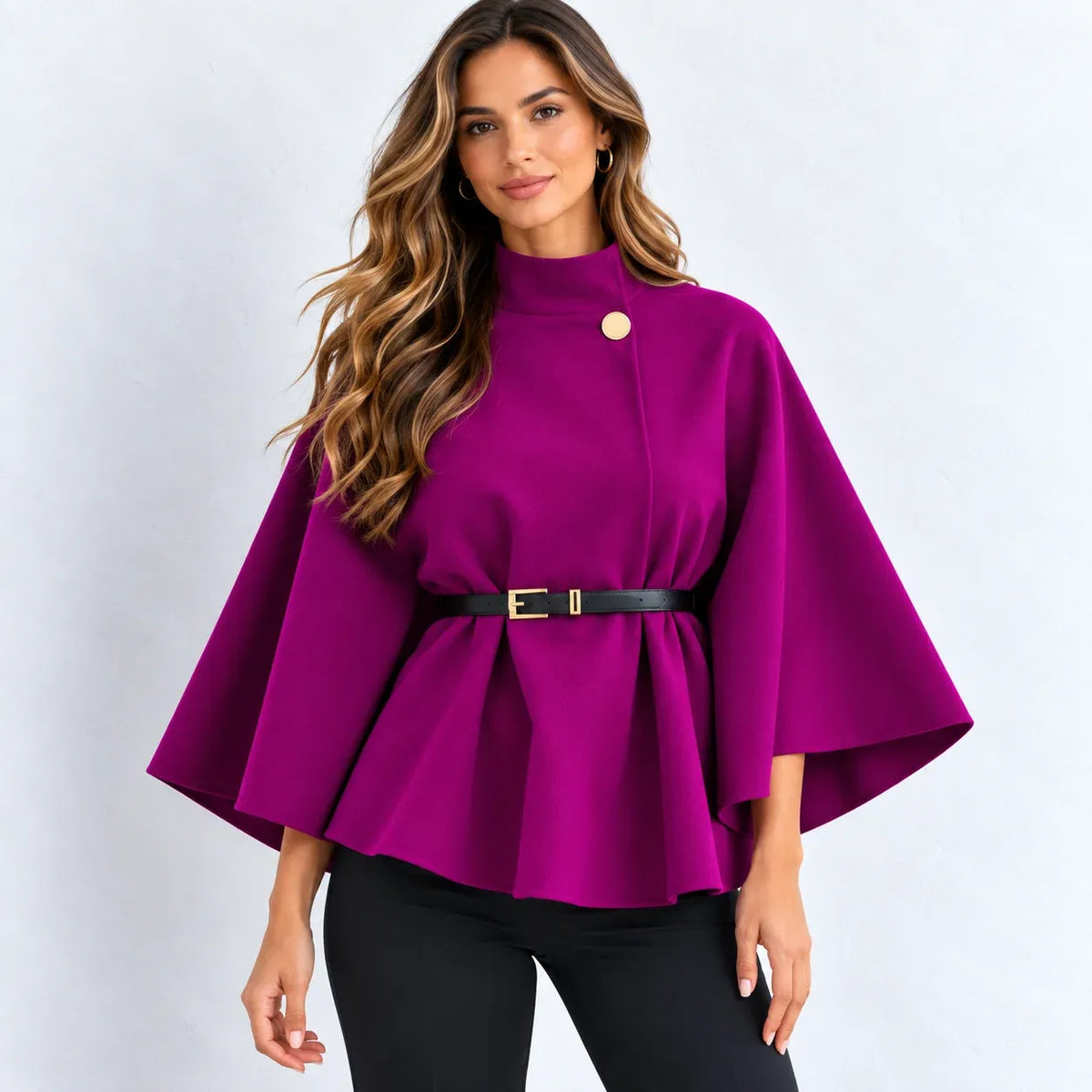 A woman modeling a bright magenta-purple cape-style top with a high mock neck, wide three-quarter sleeves, a peplum hem, a single decorative gold button, and a black belt with a gold buckle at the waist, paired with black pants against a light gray studio background.