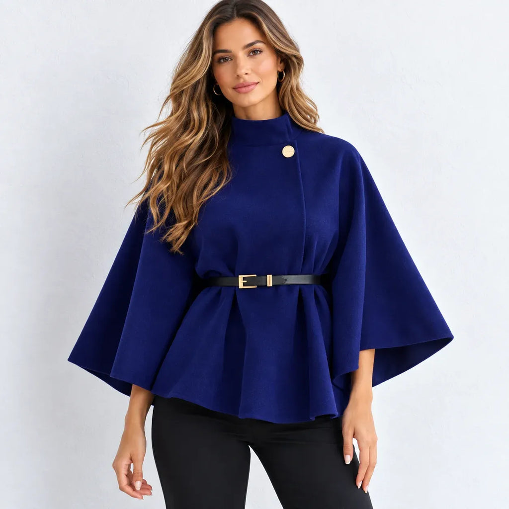 A woman modeling a royal blue cape-style peplum top with a single gold button, a black belt with a gold buckle at the waist, and black pants, set against a plain light gray studio background.